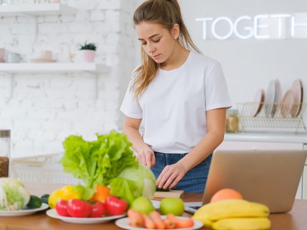 Woman in white t-shirt preparing fresh vegetables in a bright kitchen with a laptop.