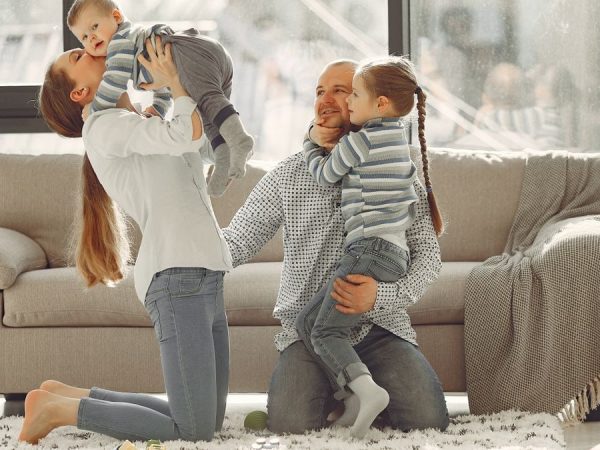 Cheerful mother kissing little boy while father hugging adorable daughter when family sitting on floor in rainy day against window